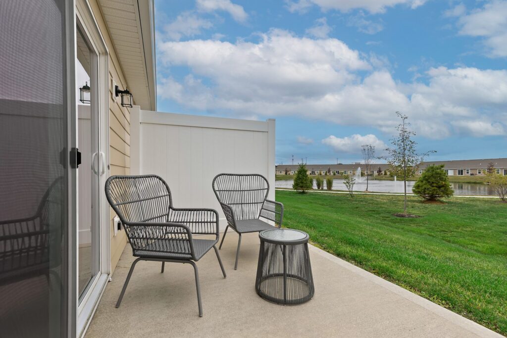 Three black metal chairs and a matching table sit on a concrete patio facing a grassy yard, small trees, and a pond under a partly cloudy sky.