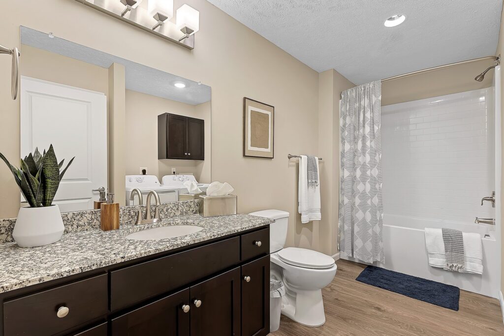 Modern bathroom with granite countertop, dark wood cabinets, toilet, bathtub with white tile, shower curtain, and laundry machines in the background.