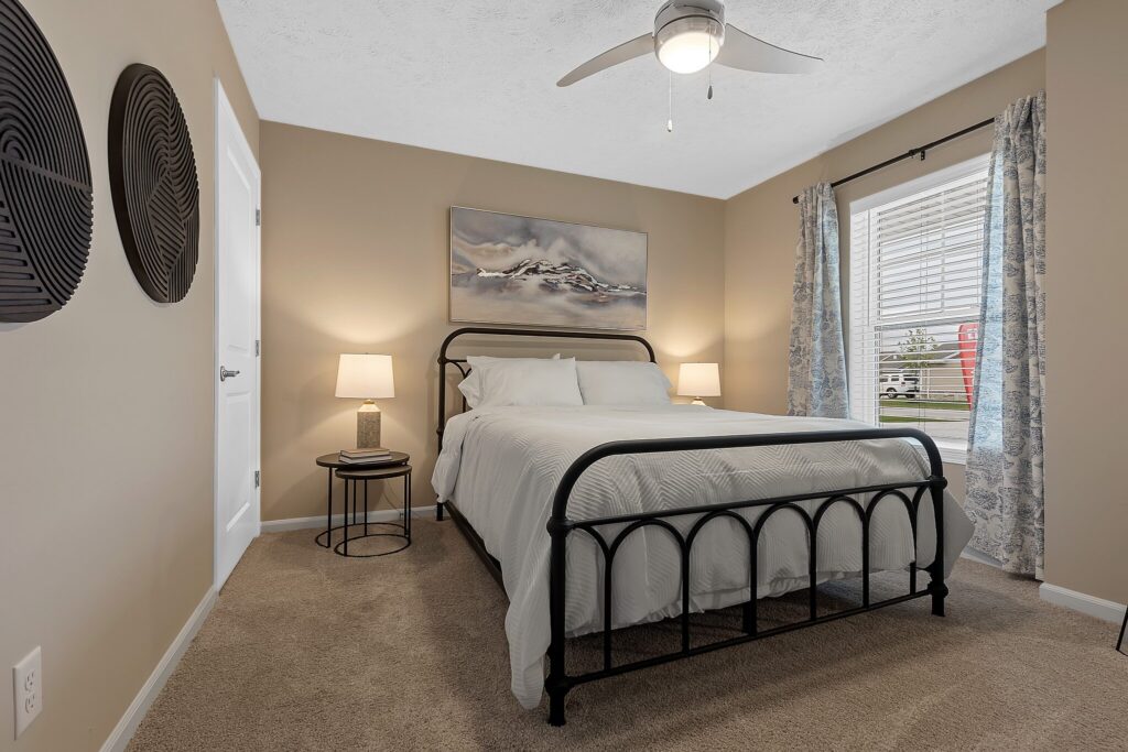 A bedroom with a black metal bed frame, white bedding, two bedside tables with lamps, beige walls, wall art, and a window with blue curtains letting in daylight.