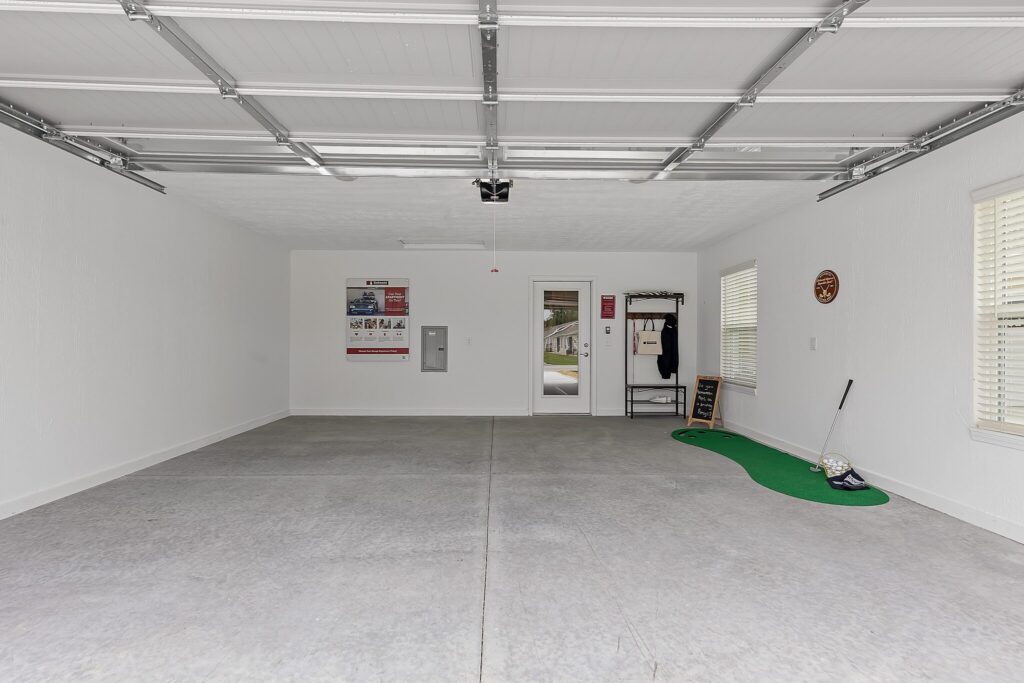Two-car garage with a concrete floor, white walls, wall posters, a door, windows, and a small putting green with golf balls and a club by the side wall.