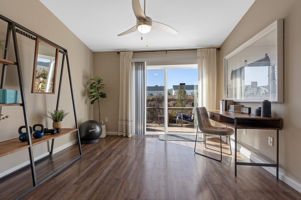 A modern home office with a desk, chair, and shelves, featuring wood flooring, large window with curtains, and a view of the outdoors.