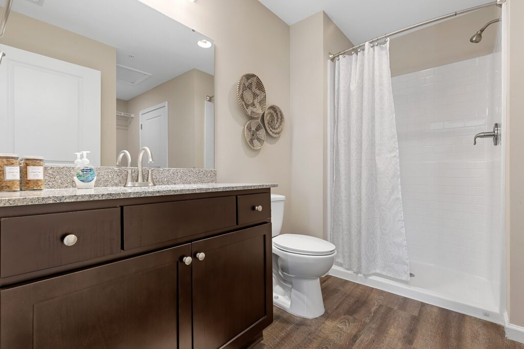 Modern bathroom with a dark wood vanity, granite countertop, toilet, and a walk-in shower with a white curtain. Three decorative baskets hang on the beige wall above the toilet.