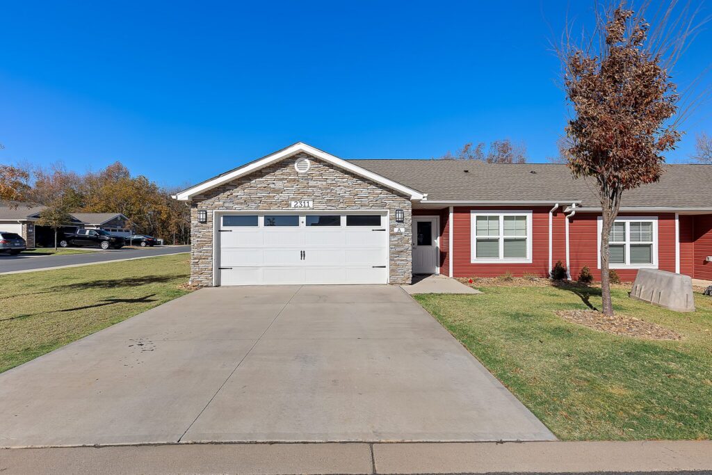 Single-story house with red siding, stone accents, a white double garage door, driveway, small tree, and neatly maintained lawn under a clear blue sky.
