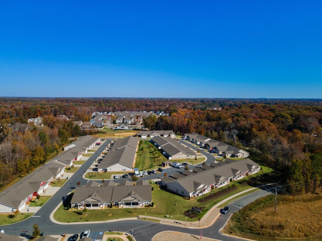 Aerial view of a suburban neighborhood with rows of houses, green lawns, surrounding roads, and a wooded area in the background under a clear blue sky.