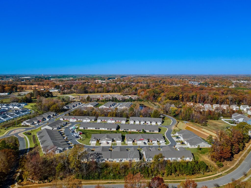 Aerial view of a suburban residential neighborhood with rows of houses, surrounded by trees with autumn foliage and open green spaces under a clear blue sky.