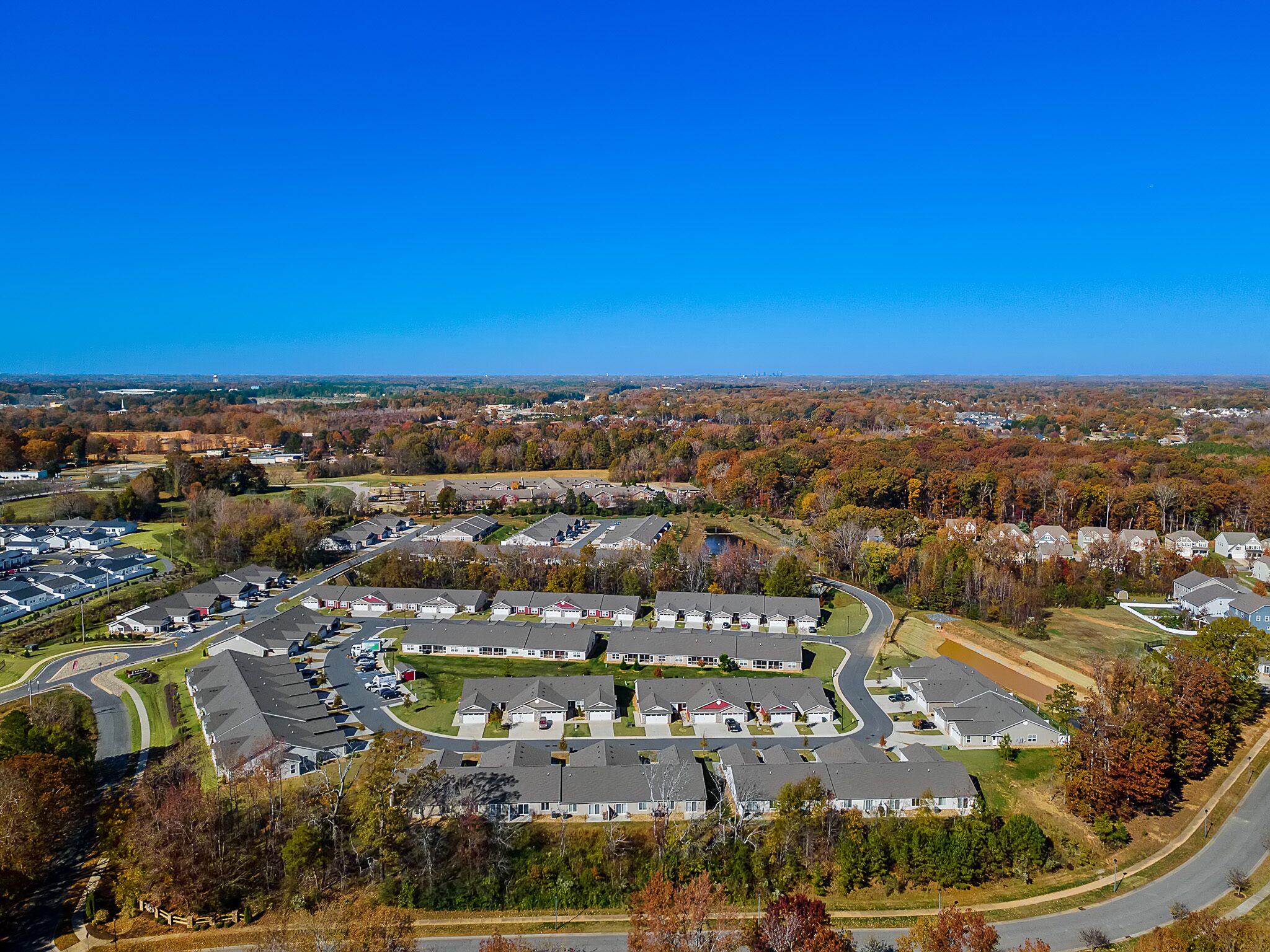 Aerial view of a suburban residential neighborhood with rows of houses, surrounded by trees with autumn foliage and open green spaces under a clear blue sky.