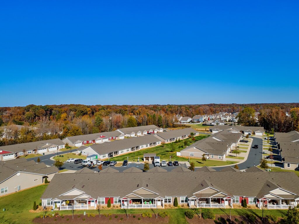 Aerial view of a suburban residential neighborhood with single-story homes, green lawns, curved streets, and a tree line in the background under a clear blue sky.