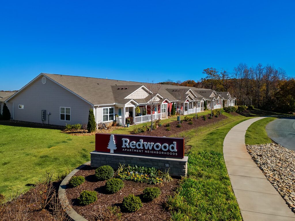 A row of single-story apartment units with manicured lawns and a sign in front reading "Redwood Apartment Neighborhoods" under a clear blue sky.