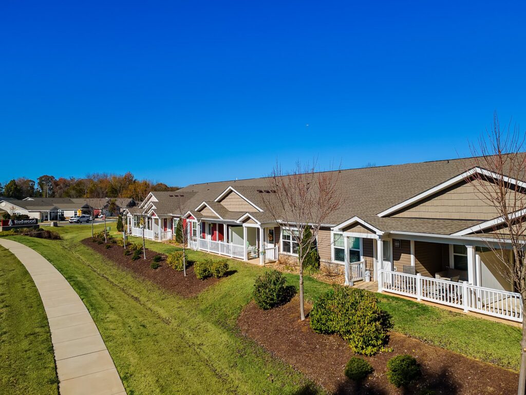 Row of single-story townhomes with front porches, landscaped lawns, and a sidewalk under a clear blue sky.