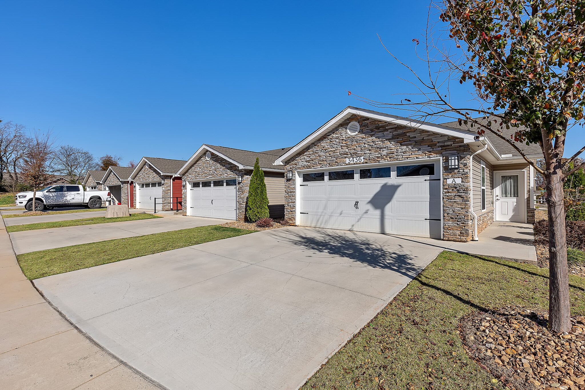 Row of modern single-story houses with stone facades, double garages, and neatly maintained lawns on a sunny day.