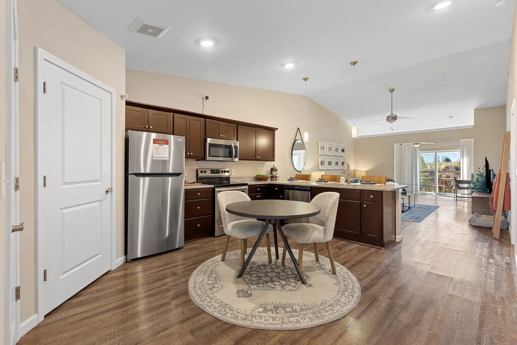 Open kitchen and dining area with dark cabinets, stainless steel appliances, round table with four chairs on a rug, and a view into the living room and balcony.