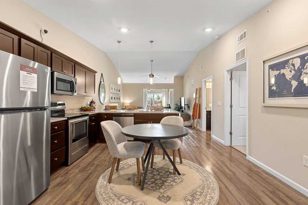 Open-concept kitchen and dining area with stainless steel appliances, dark wood cabinets, a round table with two chairs, and a living space visible in the background.