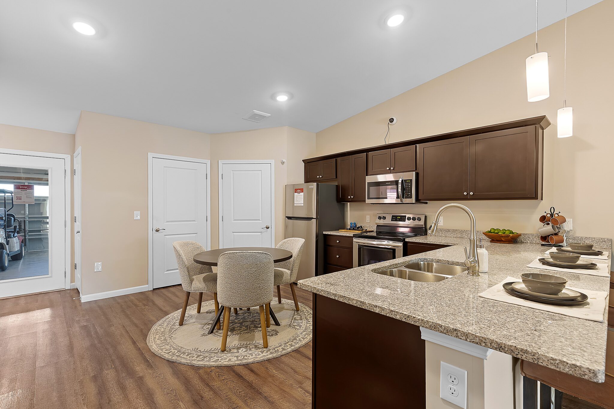 Modern kitchen and dining area with brown cabinets, stainless steel appliances, granite countertops, and a round table with four chairs on a rug.