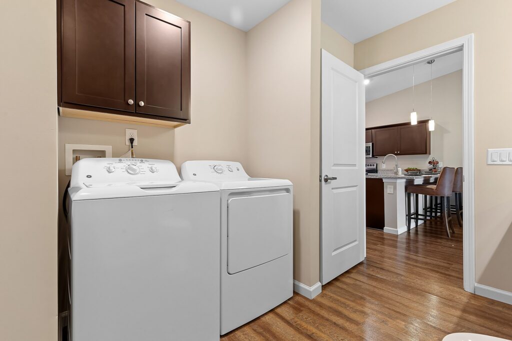 A laundry area with a white washing machine and dryer, brown cabinets above, and an open door leading to a kitchen with wood floors and pendant lights.