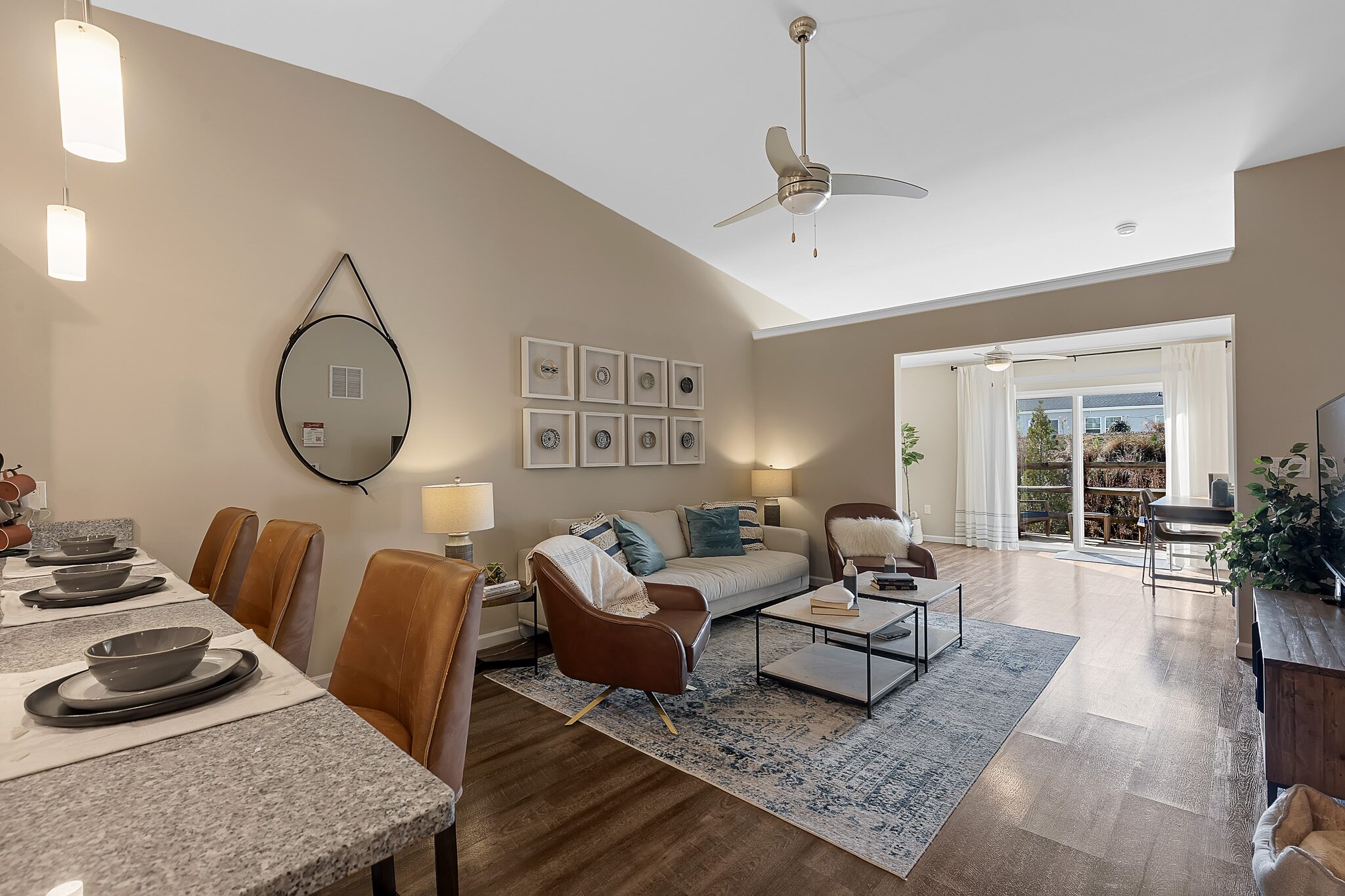 Modern living room with a neutral color palette, featuring a sofa, coffee table, TV, wall art, and a bar counter with stools, leading to a sunroom with large windows.