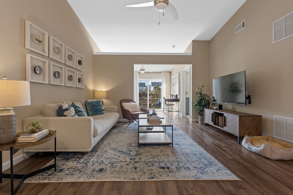 Modern living room with beige walls, a light sofa, armchair, coffee table, TV on a stand, patterned rug, and wall art. Large windows offer natural light and a view into a dining area.