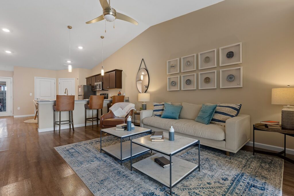 Modern living room with a beige sofa, blue accents, two-tier coffee table, kitchen with bar stools, and wall art, all set on wood flooring and a patterned area rug.