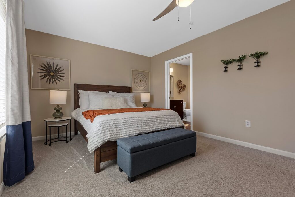 A neatly made bed with white bedding and an orange throw blanket is centered in a beige bedroom, featuring two nightstands, decorative wall art, and an upholstered bench at the foot of the bed.