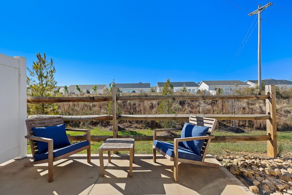 Two wooden chairs with blue cushions and a small table sit on a patio near a wooden fence, with grass, rocks, and houses visible in the background under a clear blue sky.