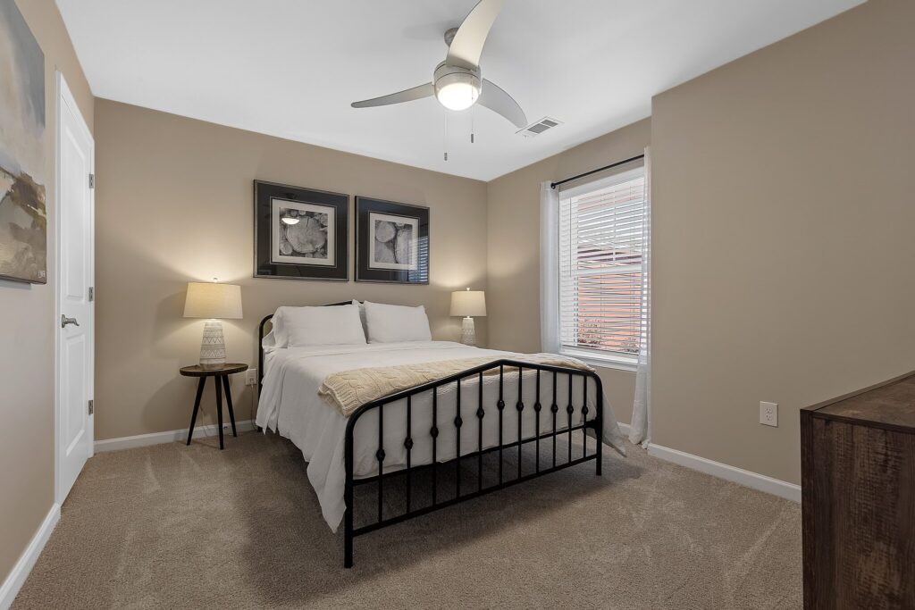 A small bedroom with beige walls, a black metal bed frame, white bedding, two bedside lamps, wall art above the bed, and a window with blinds.