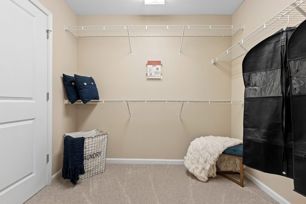 A clean walk-in closet with empty wire shelves, a laundry basket, two pillows, a black garment bag, and a bench with a white blanket.