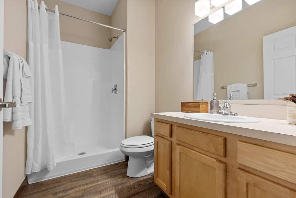 Modern bathroom with a walk-in shower, white curtain, toilet, wooden vanity with sink, mirror, and overhead lighting. Towels and soap dispenser are visible.