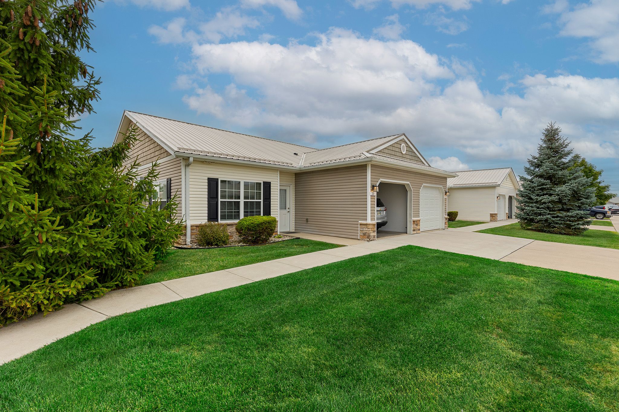 Single-story beige house with a white garage door, well-maintained lawn, and sidewalk, under a partly cloudy sky.