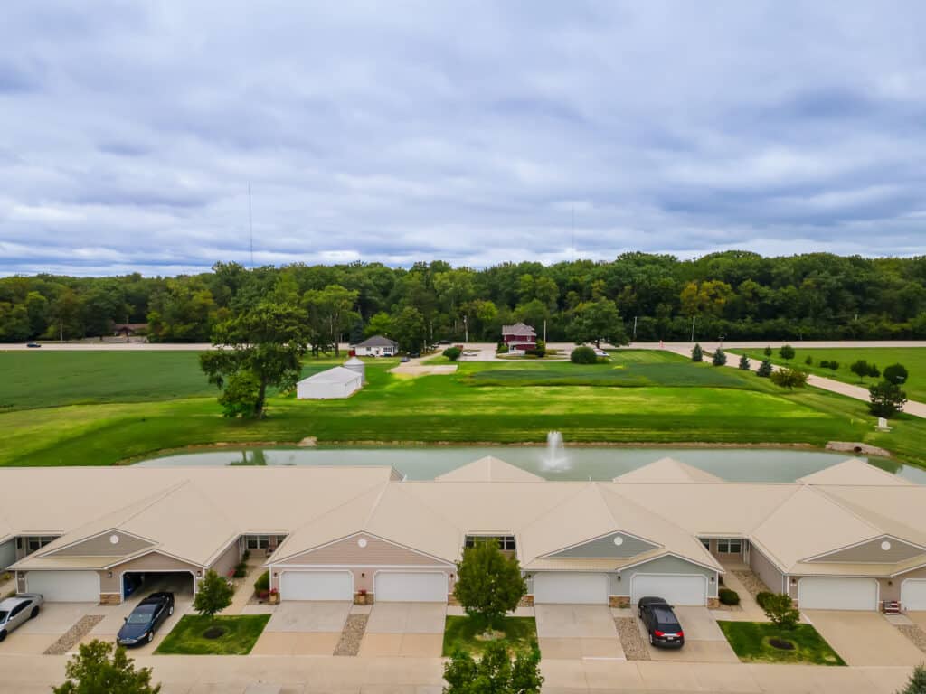 A row of townhomes with garages faces a small pond with a fountain, surrounded by green lawns, trees, and a few distant houses under a cloudy sky.