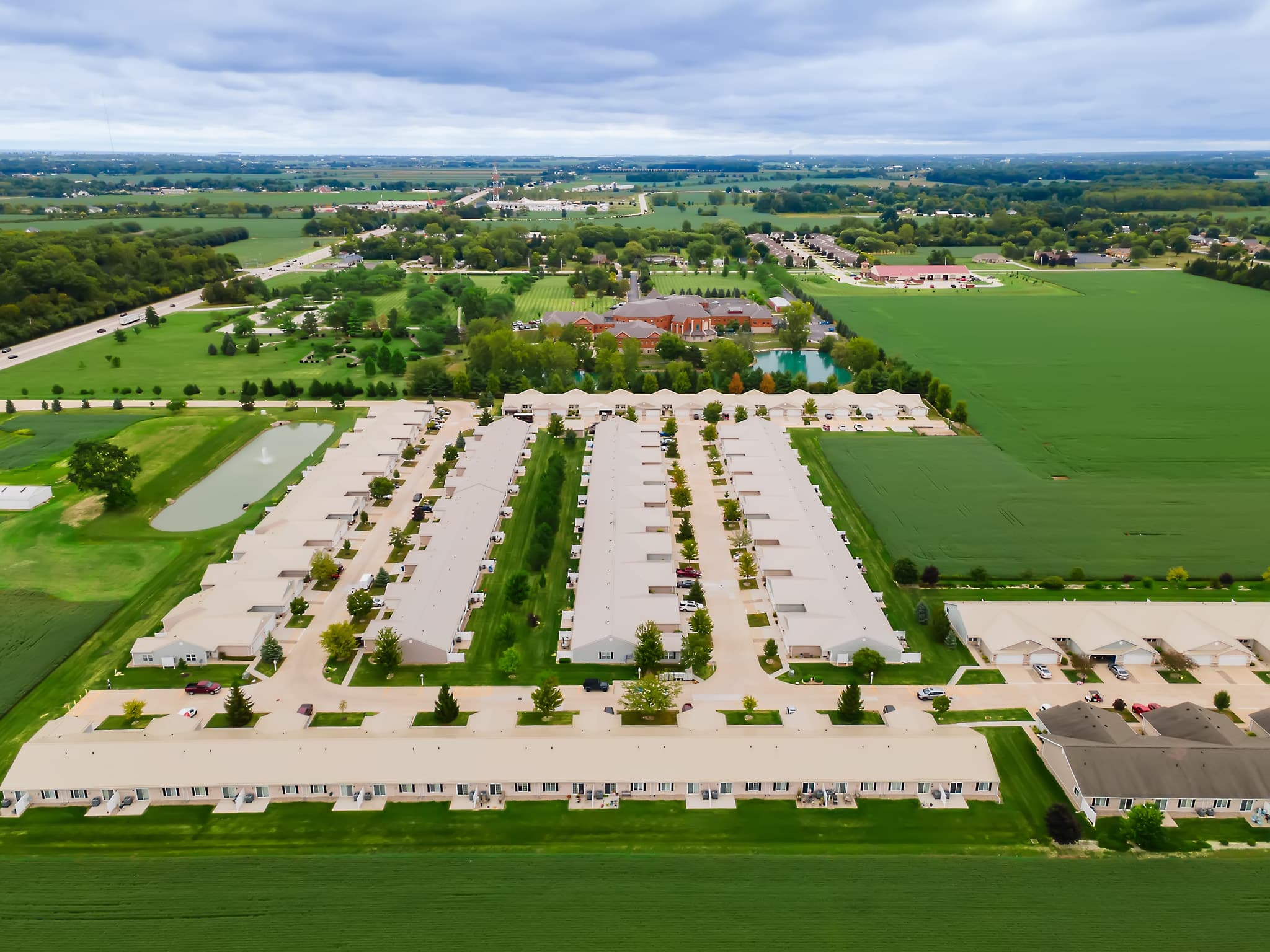 Aerial view of a residential neighborhood with rows of identical single-story houses surrounded by green fields and roads under a cloudy sky.