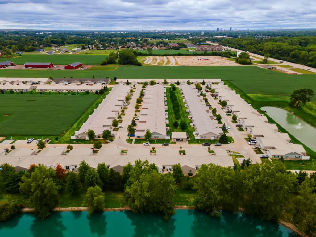 Aerial view of a suburban residential neighborhood with rows of houses, green fields, trees, a pond in the foreground, and a city skyline in the distant background under a cloudy sky.