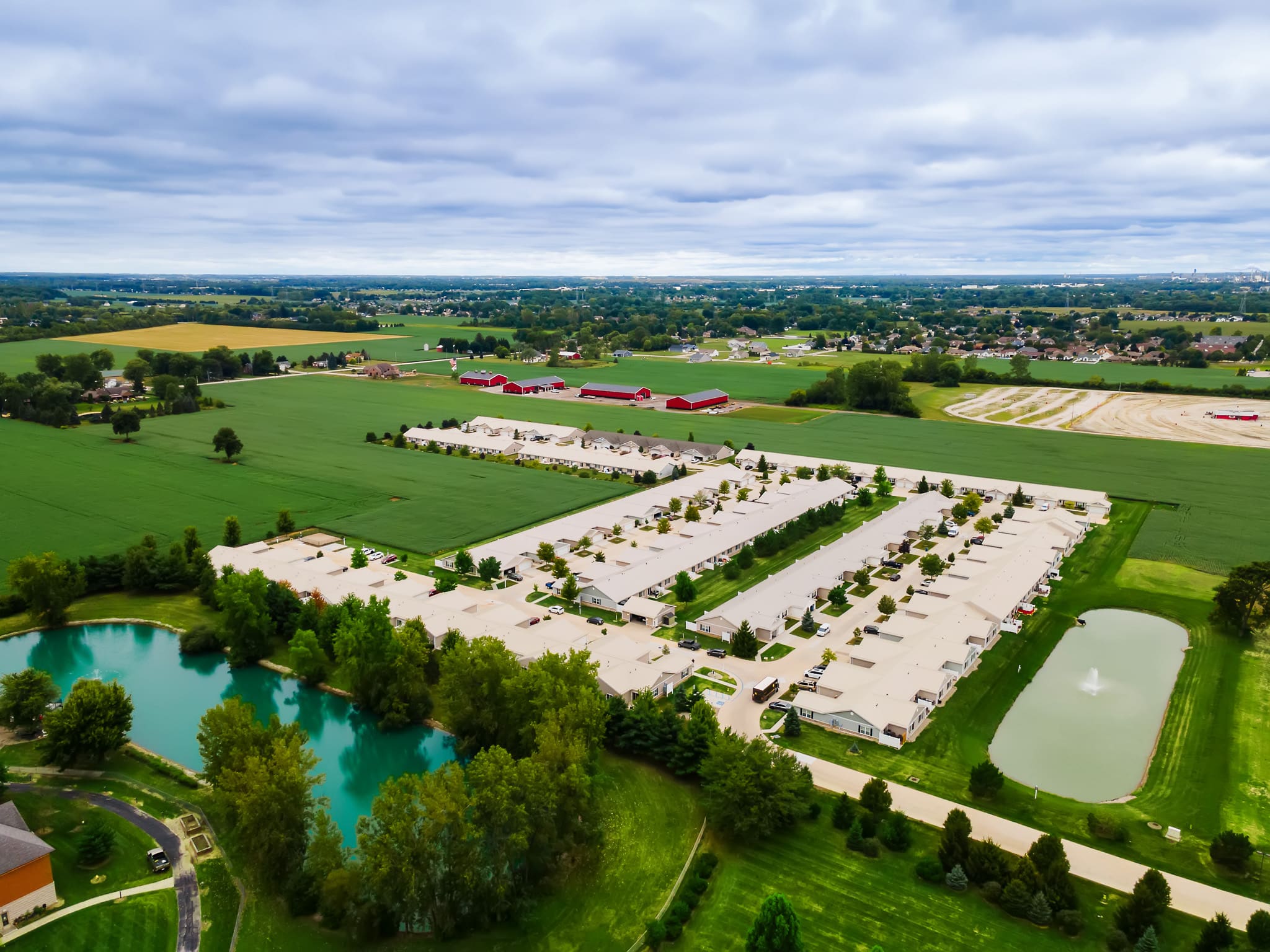 Aerial view of a residential community with rows of white-roofed houses, surrounded by green fields, trees, ponds, and distant farmland under a cloudy sky.