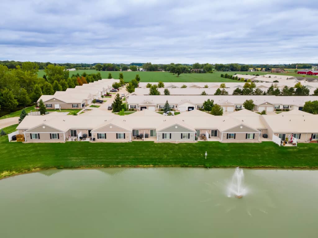 Aerial view of single-story residential buildings arranged in rows near a pond with a water fountain, surrounded by green lawns and trees under a cloudy sky.