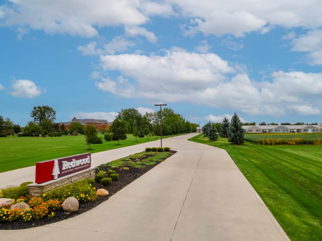 A wide concrete driveway leads through a landscaped lawn with a "Redwood" sign on the left and buildings in the distance under a partly cloudy sky.
