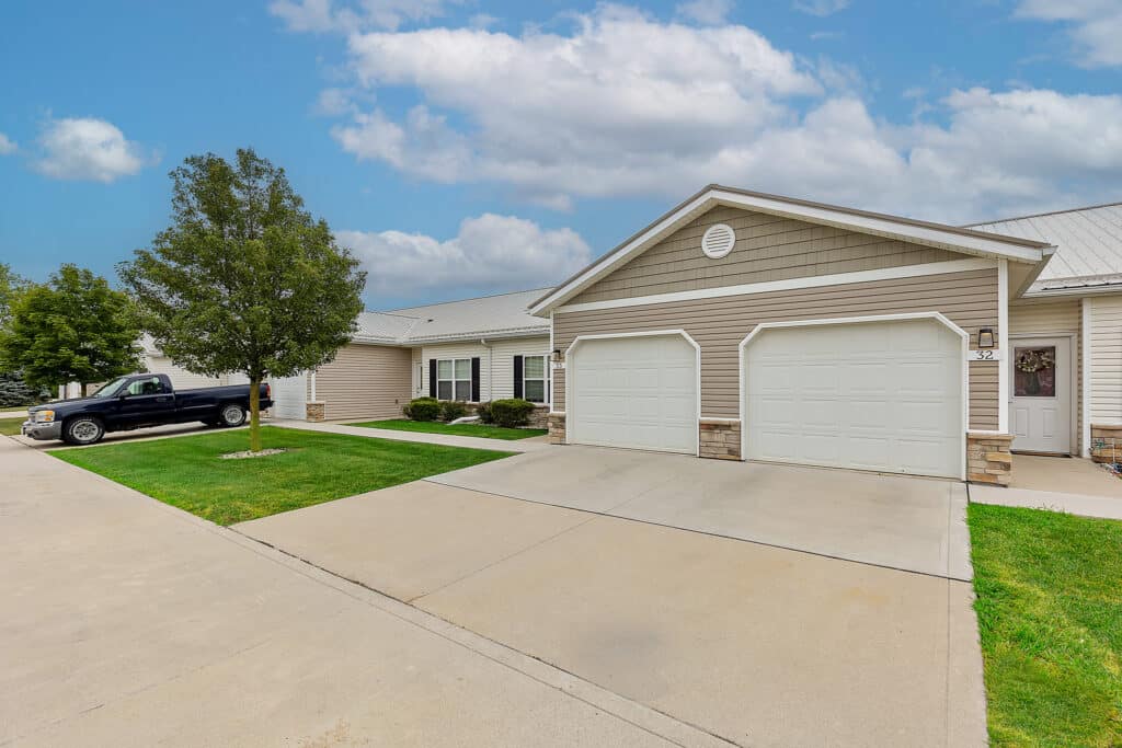 Single-story beige house with two white garage doors, a pickup truck parked in the driveway, and a well-maintained lawn under a partly cloudy sky.