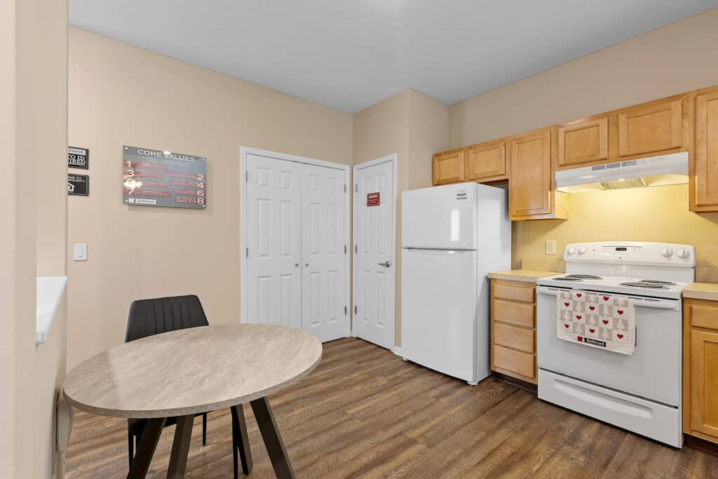 A small kitchen with light wood cabinets, white appliances, a round table with two chairs, and a sign with core values on the wall.