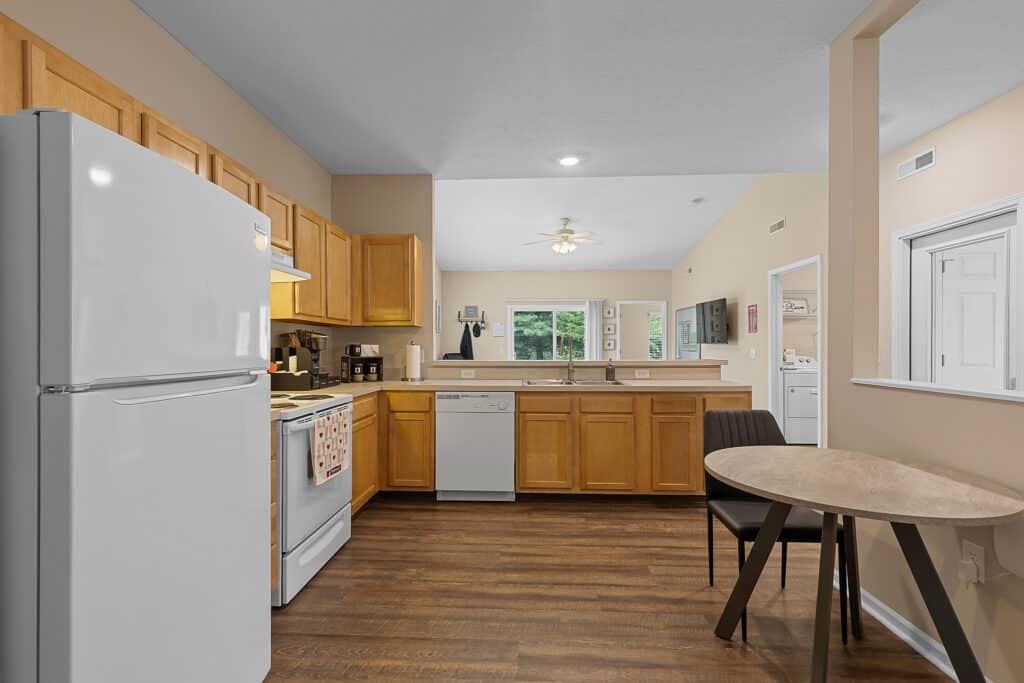 Modern kitchen with light wood cabinets, white appliances, dishwasher, and round dining table with two chairs on a wood floor. Large window provides natural light.