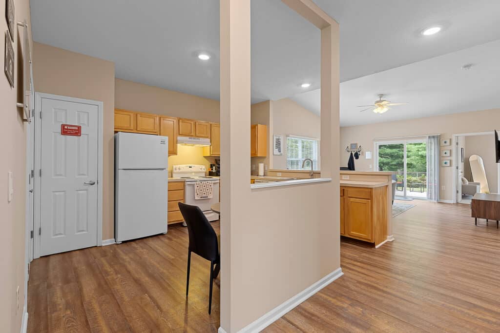 Open kitchen and dining area with wood flooring, light wood cabinets, white appliances, and a view of a living room leading to a patio with glass doors.