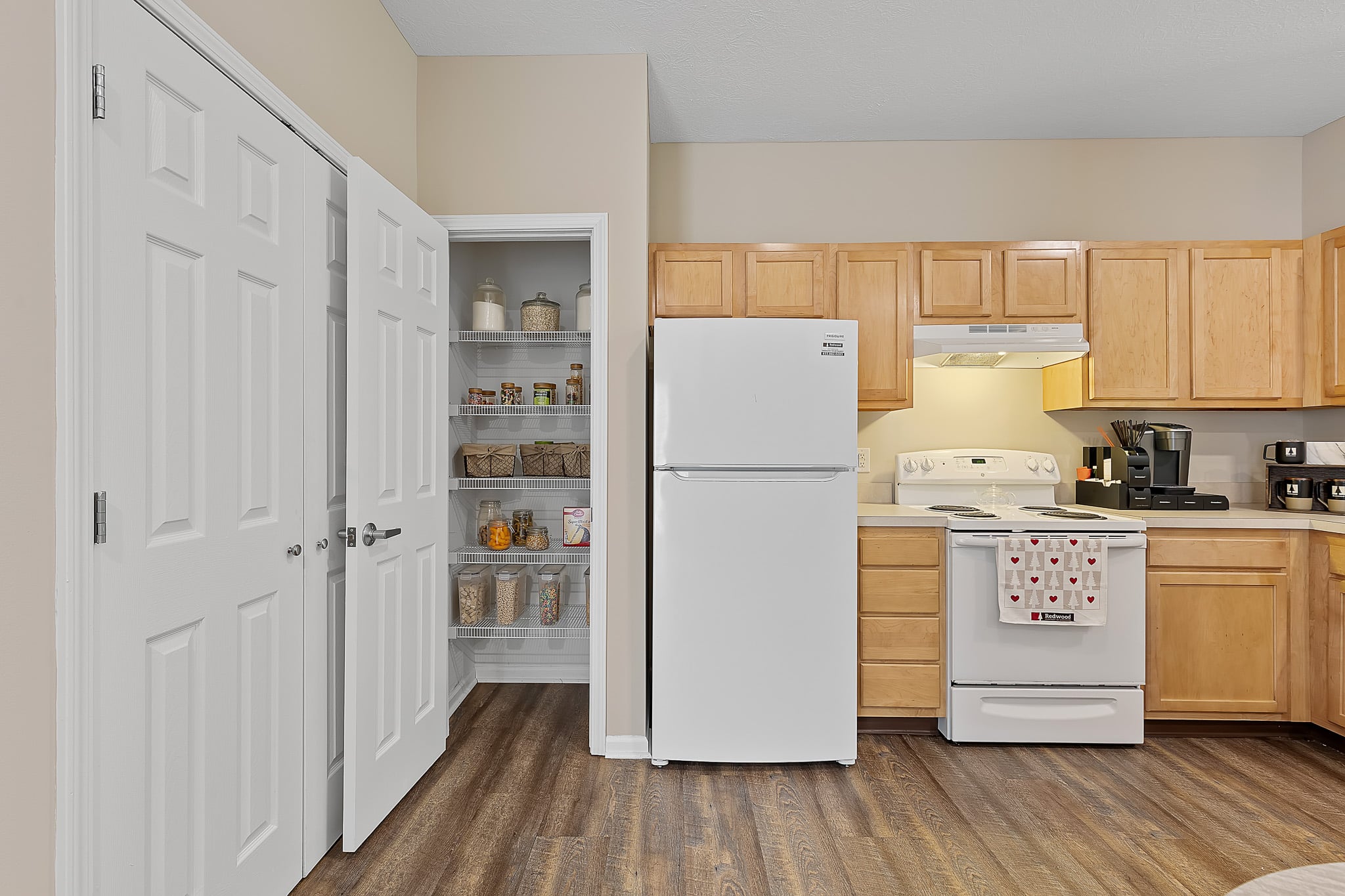 A kitchen with light wood cabinets, white appliances, open pantry shelves stocked with jars, and a wood floor.