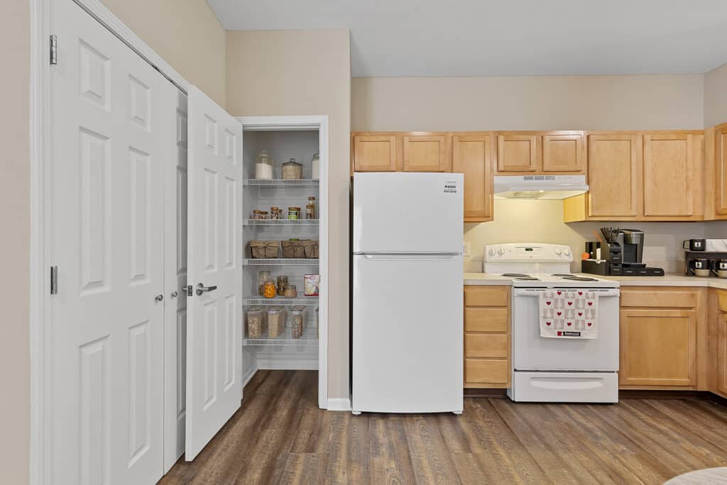 A kitchen with light wood cabinets, white appliances, a double-door pantry with shelves of jars, and wood-style flooring.