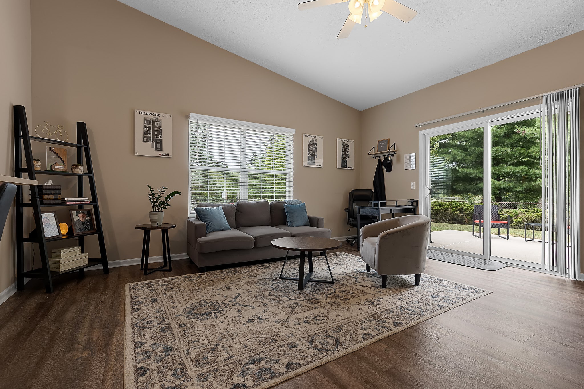 A modern living room with a gray sofa, armchair, round coffee tables, large rug, bookshelf, and glass sliding doors leading to a patio with greenery outside.