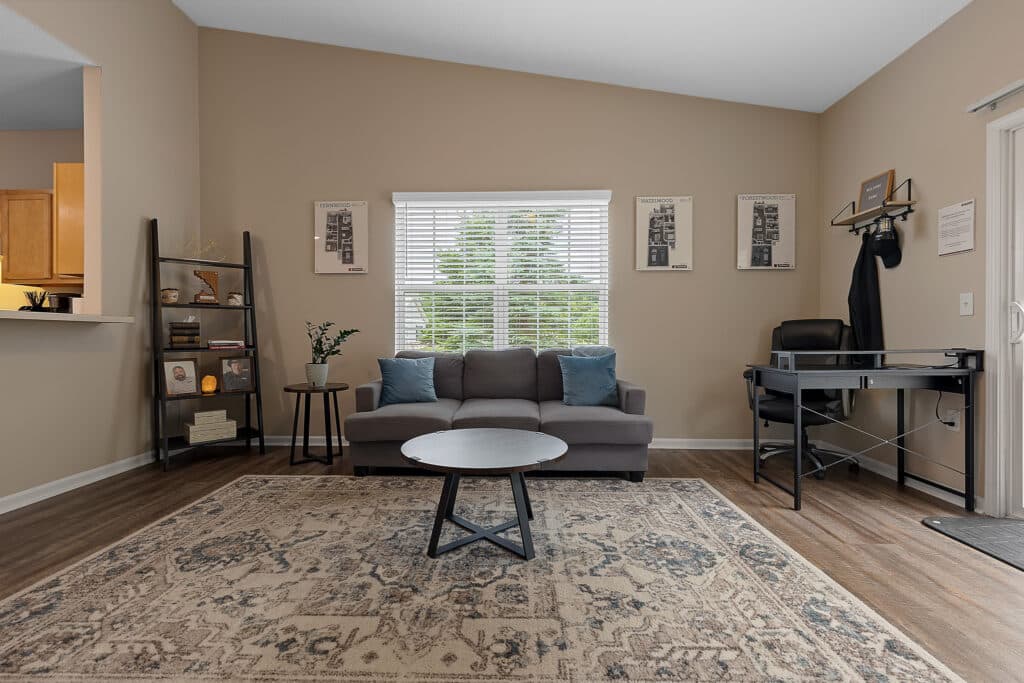 A living room with a gray sofa, a round coffee table, a patterned rug, a black bookshelf, and a small desk by the window with blinds. Walls are beige and floor is wood.