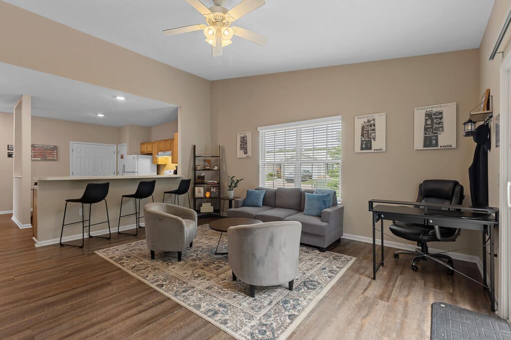 Modern living room with a gray sofa, two chairs, a patterned rug, a bar with stools, a small office desk, and wood flooring. Light pours in from a window behind the sofa.