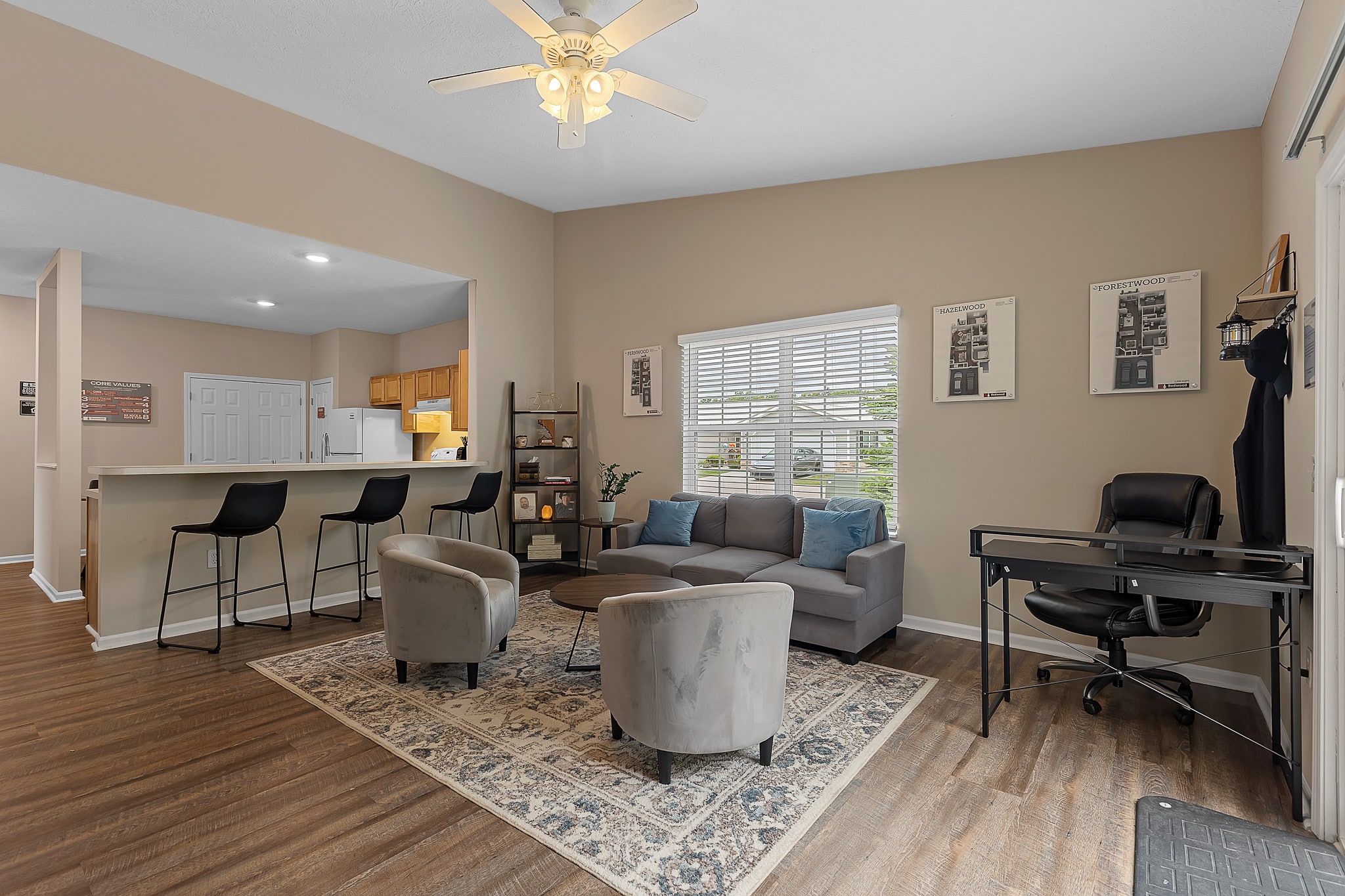 Modern living room with a gray sofa, two chairs, a patterned rug, a bar with stools, a small office desk, and wood flooring. Light pours in from a window behind the sofa.