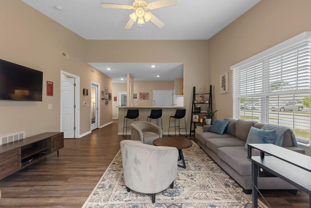 Modern living room with neutral decor, featuring a sofa, armchairs, coffee table, TV, bar stools at a kitchen counter, large window, and wood flooring.