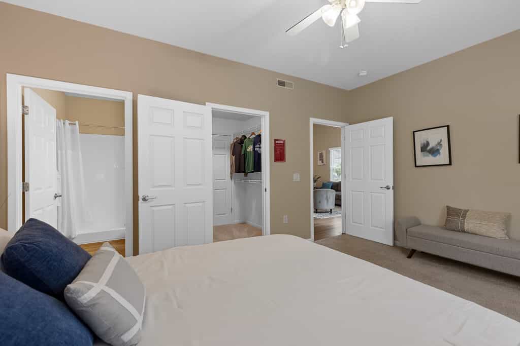 Bedroom with a neatly made bed, open doors to a walk-in closet and bathroom, beige walls, ceiling fan, and a gray bench along one wall.