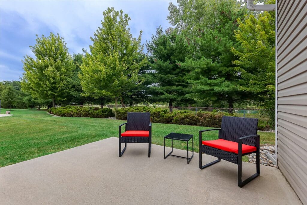 Two black patio chairs with red cushions and a small black table sit on a concrete patio, overlooking a grassy yard with trees and a chain-link fence.