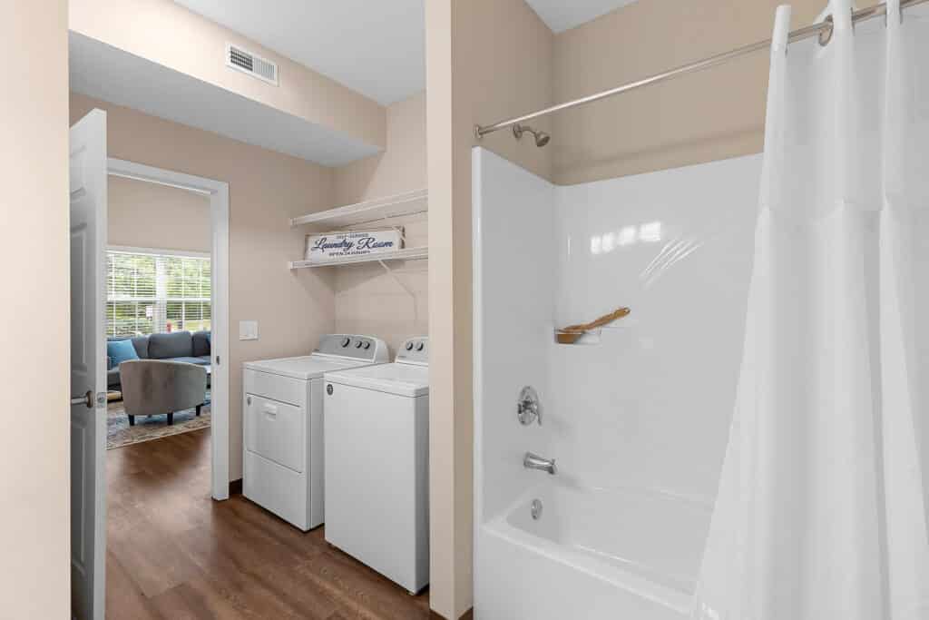 Laundry area with washing machine and dryer next to a bathroom with a white bathtub and shower curtain; living room visible through open door.