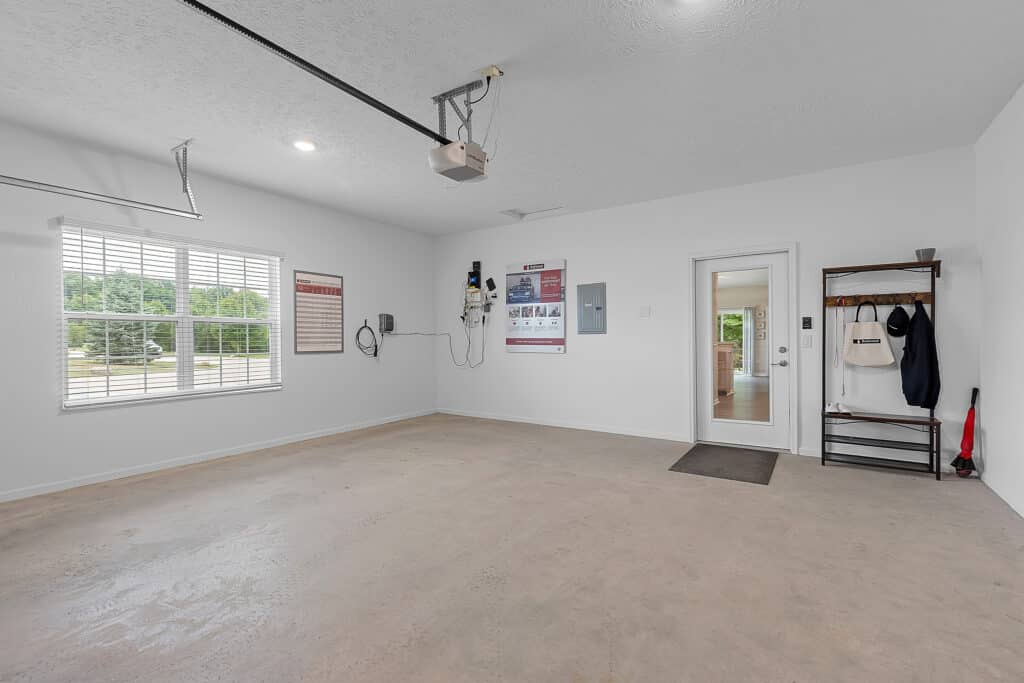 Clean, empty garage with a window, wall-mounted equipment, a door to the interior, a small entryway mat, and an organized coat rack with hanging items by the door.