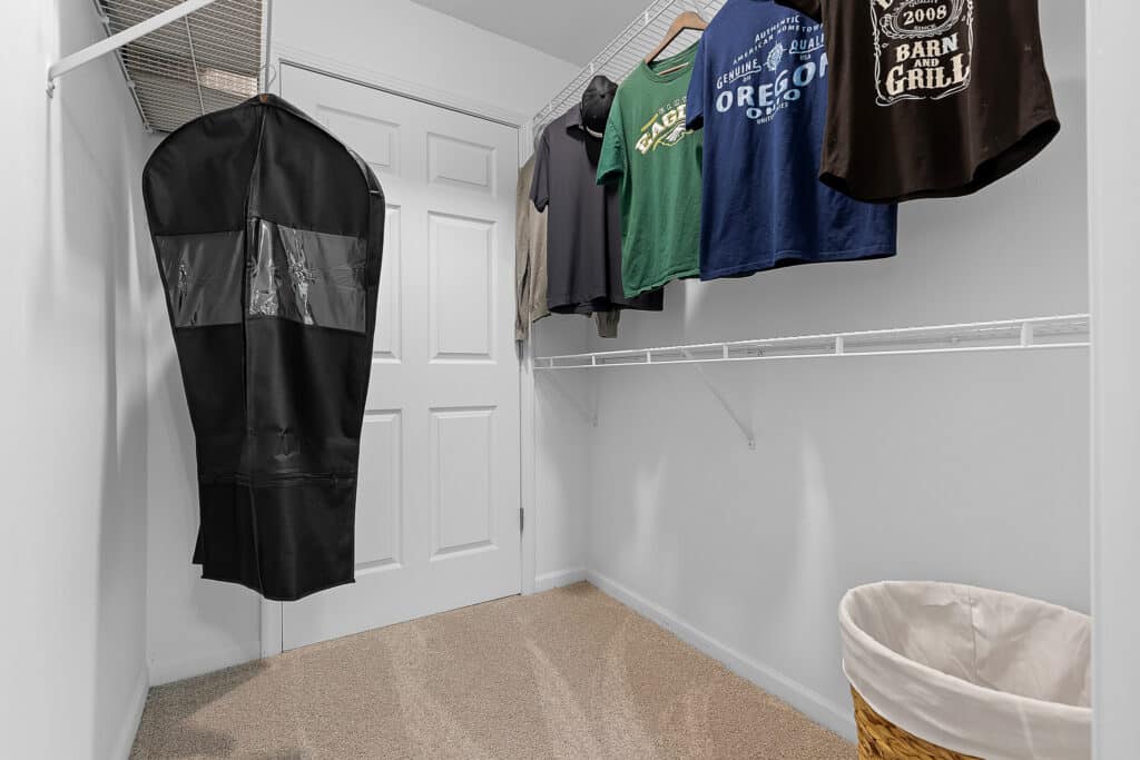 A walk-in closet with a few shirts hanging on wire racks, a garment bag, and a laundry basket on a beige carpeted floor.