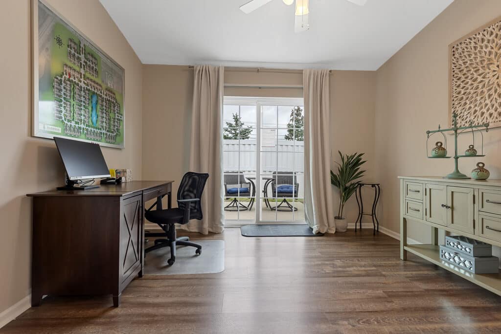 Home office with a desk, computer, and chair on the left, light green cabinet and decor on the right, plant in the corner, and glass doors leading to a patio with outdoor seating.